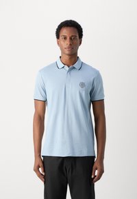Polo de algodón azul claro con cuello, manga corta y acentos negros. Presenta un logotipo bordado en el pecho.