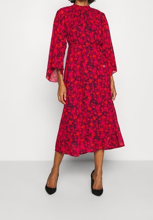 Mujer con un vestido midi rojo con estampado floral, mangas largas acampanadas y tacones negros, de pie frente a un fondo blanco liso.