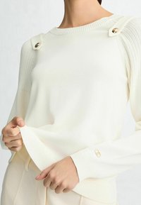 Slonovinový pletený svetr se žebrovaným vzorem na ramenou a zlatými knoflíkovými detaily. Má kulatý výstřih a žebrované manžety, což vytváří hladkou texturu.