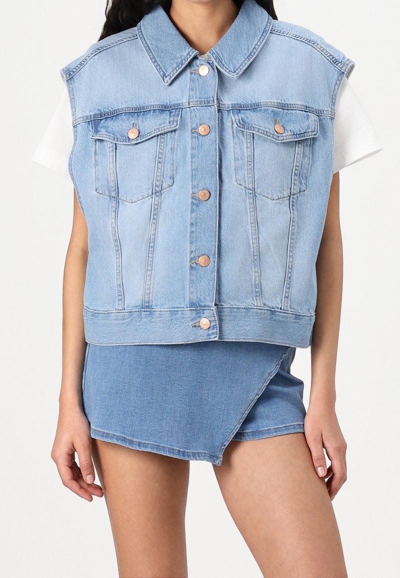 Light blue denim sleeveless jacket with a collar, front pockets, and copper buttons; paired with a matching denim wrap skirt.