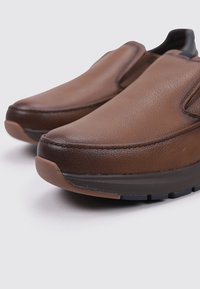 Zapatos marrones tipo mocasín hechos de cuero texturizado. Tienen la punta redonda y una suela gruesa con un acento oscuro contrastante en la parte inferior.