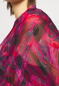 Blusa trasparente in vivaci tonalità di rosa e rosso, con un motivo floreale arricchito da perline di colore viola scuro e nero. Tessuto leggero, design fluido.
