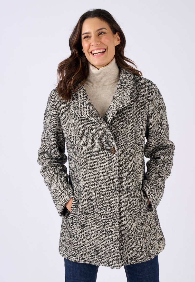 Manteau en mélange de laine noir et blanc texturé avec col à revers, fermeture à bouton unique et poches latérales, porté sur un col roulé beige.