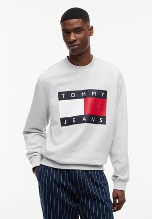 Mężczyzna w jasno szarej bluzie z dużym logo Tommy Jeans w kolorach białym, czerwonym i czarnym, w połączeniu z granatowymi spodniami w prążki.