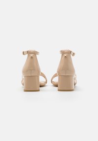 Beige patentlær heelskinnsandaler med en kraftig hæl, ankelrem og detaljer i gull, som viser jevn tekstur og design med åpen bak.