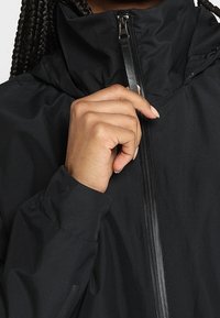 Personne avec des cheveux tressés tenant la fermeture éclair d'une veste noire près du col, se préparant à la fermer.