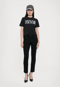 Juoda marškinėliai su baltu "VERSACE JEANS COUTURE" logotipu, derinami su juodomis siaurėjančiomis kelnėmis ir pilku kepurėliu. Modelis vilki juodus aukštakulnius batus.