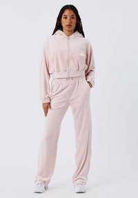 Tuta in velour rosa chiaro composto da giacca con cappuccio e zip, dotata di tasche frontali, e pantaloni con vita regolabile e vestibilità rilassata.