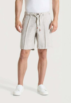 Uomo che indossa pantaloncini di lino beige con coulisse e sneakers bianche, in piedi davanti a uno sfondo neutro.