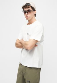 Νέος άνδρας που φοράει λευκό t-shirt, χακί πράσινα σορτς με λογότυπο, μπεζ καπέλο φοράει ανάποδα και μαύρα γυαλιά ηλίου τύπου wraparound, με σταυρωμένα τα χέρια.