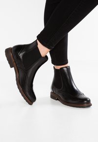 Stivali Chelsea brogue in pelle nera con pannelli elastici laterali indossati con pantaloni neri su uno sfondo bianco.