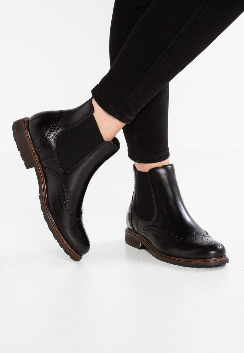 Stivali Chelsea brogue in pelle nera con pannelli elastici laterali indossati con pantaloni neri su uno sfondo bianco.