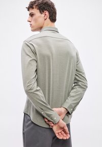 Majica z dolgimi rokavi v svetlo zeleni barvi, izdelana iz teksturirane tkanine, z ovratnikom in manšetami na gumbe ter ukrivljenim robom in detajli na zadnjem šivu.