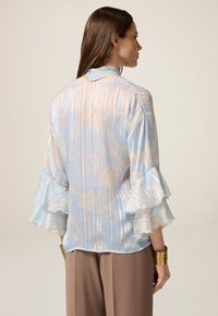 Blusa azzurro chiaro con maniche a ruffle, caratterizzata da un sottile motivo a righe e un design astratto in tonalità beige. Tessuto leggero e fluido.