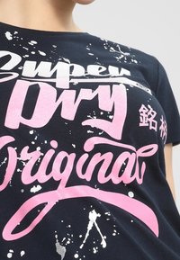 Granatowa koszulka bawełniana z białym i różowym nadrukiem graficznym z efektem plam. Tekst zawiera "Super Dry Original" w pogrubionej czcionce.