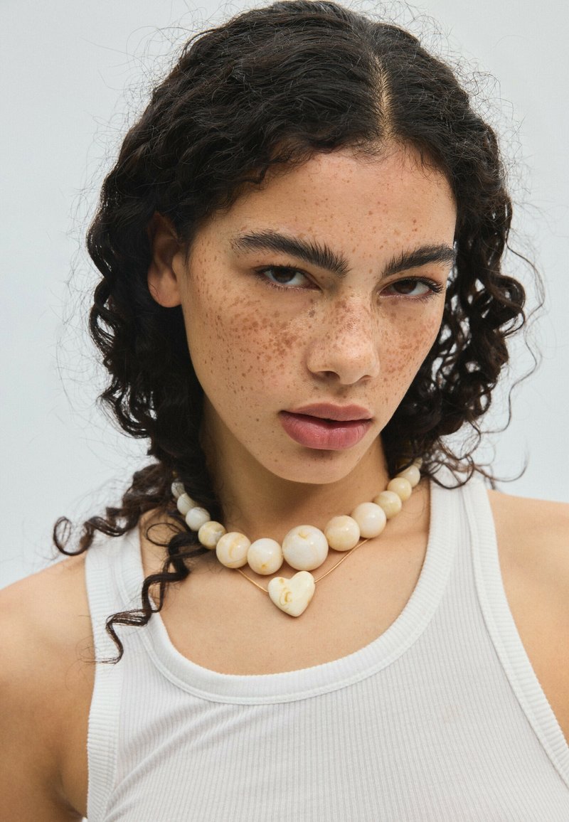 Jeune femme aux cheveux bouclés et foncés, taches de rousseur, portant un débardeur blanc et deux colliers couleur crème, un avec des perles rondes et un avec un pendentif en forme de cœur.