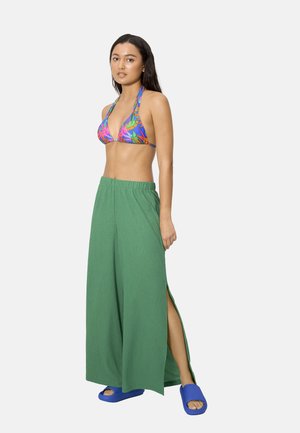 Mujer que lleva un top de bikini floral colorido, pantalones verdes anchos y sueltos con aberturas laterales, y sandalias azules tipo chancla, de pie contra un fondo liso.