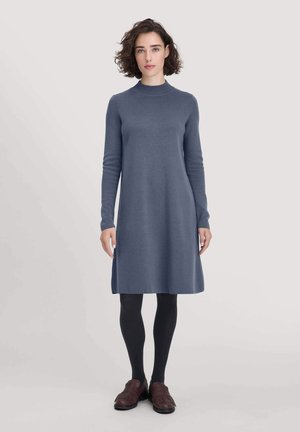 Abito in maglia in un morbido tessuto blu scuro, con maniche lunghe e colletto alto, che arriva appena sopra il ginocchio. Abbinato a collant neri e scarpe marroni.