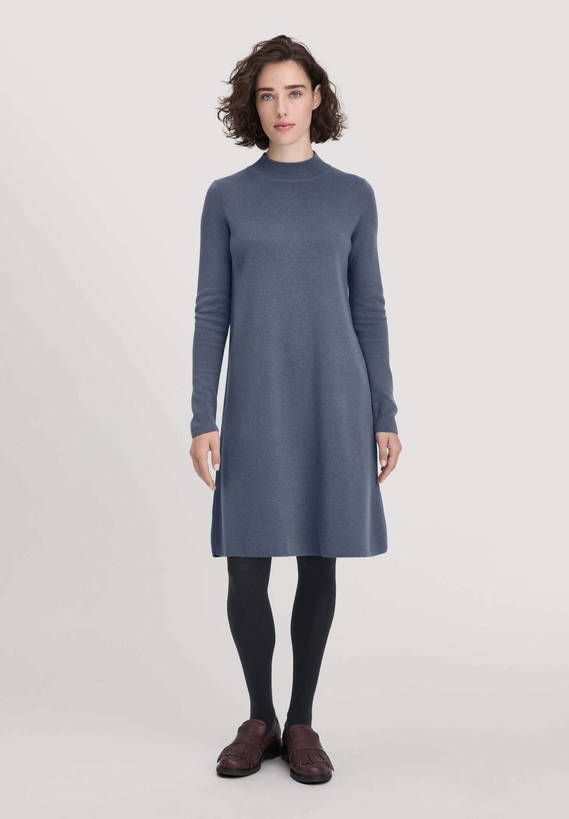 Gebreide jurk van een zachte, donkerblauwe stof met lange mouwen en een hoge halslijn, vallend net boven de knie. Gepaarde met zwarte panty’s en bruine schoenen.