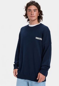 Granatowy, luźny sweter z prążkowanymi ściągaczami na mankietach i na dole, z białym napisem "OUTSIDE" na lewej części klatki piersiowej. Miękka faktura i dzianinowy wzór.