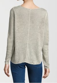 Femme portant un pull en maille à manches longues de couleur beige clair avec une couture centrale dans le dos, associé à un jean bleu foncé.