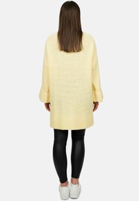 Maglione oversized giallo a maglia con maniche lunghe, texture a coste e orlo arrotondato, abbinato a leggings neri e sneakers bianche.