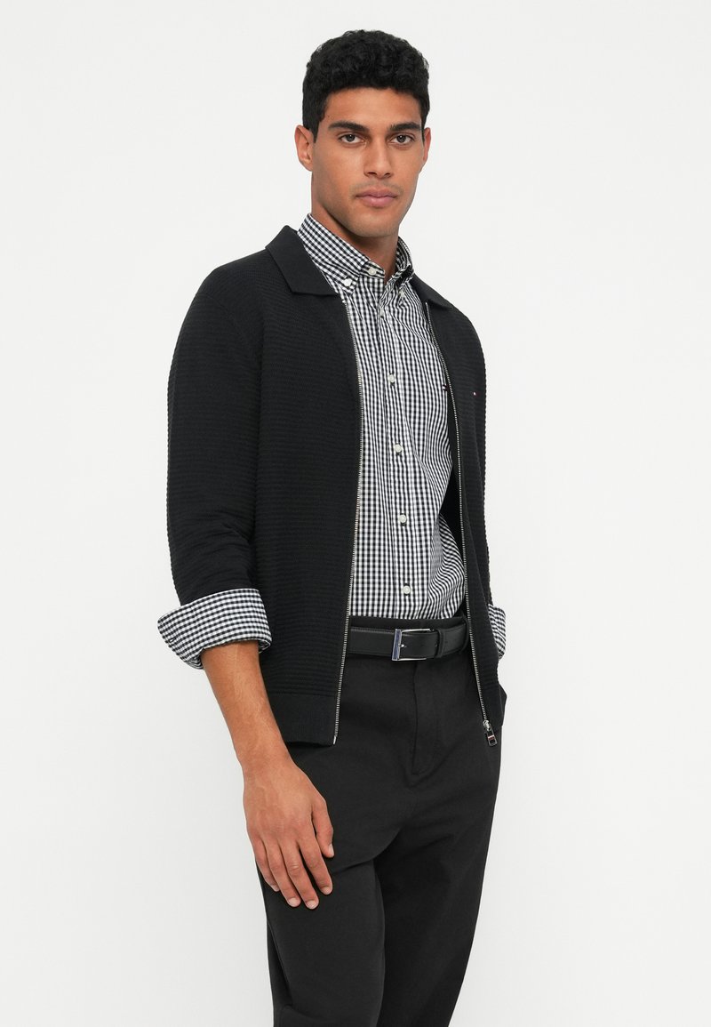 Black zip-front cardigan with ribbed texture, worn over a black and white gingham shirt, paired with black pants and a belt.