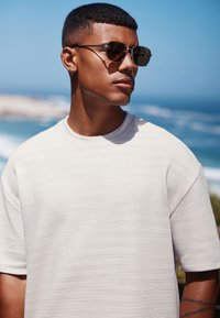 Ljusgrå ribbad t-shirt med rund halslinning, korta ärmar och slät yta. Svarta runda solglasögon, kustbakgrund med blått hav.