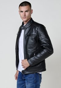 Giacca di pelle nera con colletto alto, tasche con zip e spalle a coste. Abbinata a una camicia bianca e jeans blu.