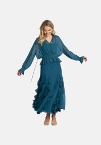 Abito maxi color turchese con maniche lunghe trasparenti e orlo arricciato. Presenta una vita aderente e una silhouette fluida, realizzato in tessuto leggero.
