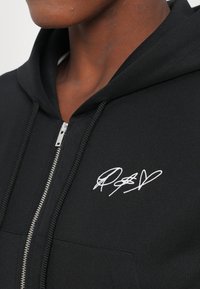Felpa nera con zip realizzata in tessuto morbido, con tasca frontale e un dettaglio di firma ricamato in bianco nella parte sinistra del petto.