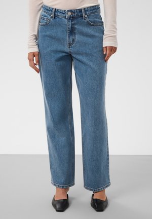 Personne portant un jean bleu droit, un haut beige à manches longues et des chaussures plates noires à bout pointu, debout sur un sol gris clair.