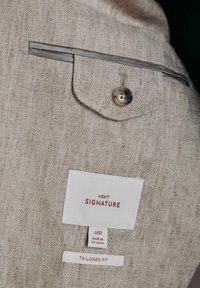 Bézs halszálka mintás szövet gombos zsebfedéllel és három ruhacímkével, melyeken a következők olvashatók: „NEXT SIGNATURE”, „40R Made in Vietnam” és „Tailored Fit”.
