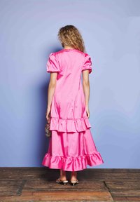 Ženska stoji ob modri steni v sijoči roza obleki s plasti in nabranimi rokavi ter drži majhno torbico iz perlic.