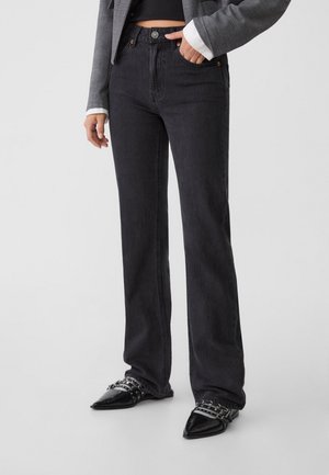 Person iført højtaljede sorte jeans med lige ben, sorte spidse sko med remme og en grå blazer med en sort top.