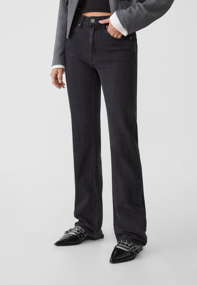 Personne portant un jean noir taille haute à jambes droites, des chaussures noires pointues avec des brides, et un blazer gris avec un haut noir.