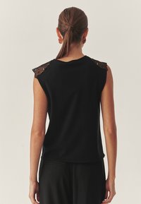 Fekete ujj nélküli felső csipkés díszítéssel a vállakon, puha textúrával, kerek nyakkivágással és laza szabással, fekete nadrággal viselve.