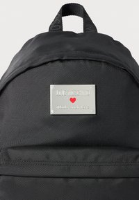 Melnā teksturētā mugursoma ar sudraba metāla plāksni, uz kuras redzams "LOVE MOSCHINO", sarkans sirds simbols un teksts "Made with love".