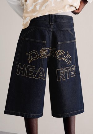 Tmavě modré džínové šortky s velkými zadními kapsami a výšivkou "DAZED HEARTS" přes zadní část, které nosí osoba s lehkým vzorovaným tričkem.
