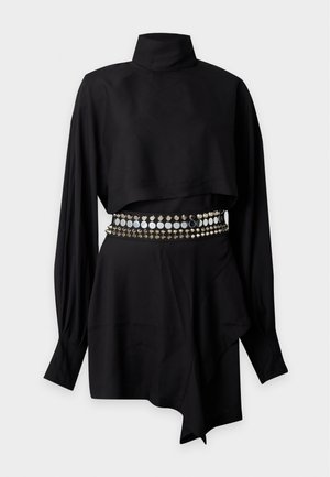 Femme portant un crop top noir à manches longues, une mini-jupe noire avec ceinture cloutée, des talons hauts noirs et des lunettes de soleil foncées sur un fond uni.