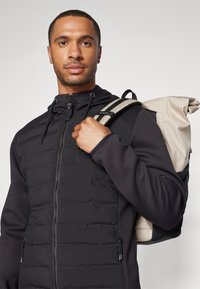 Svart zip-up jakke med polstret front, kombinert med en beige og svart ryggsekk med rull-top design og stripete stropper.