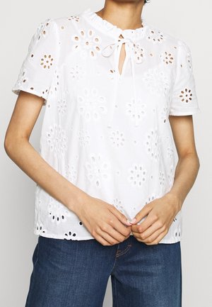 Frau trägt eine weiße Bluse mit kurzen Ärmeln, floralen Lochstickmustern und einer Krawatten-Schleifen-Verzierung am Ausschnitt, kombiniert mit blauen Jeans aus Denim.