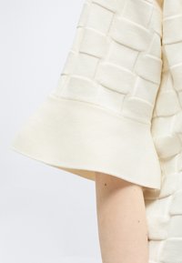 Gros plan sur une manche de couleur crème avec un motif tissé texturé et un large poignet côtelé évasé sur un bras à la peau claire.