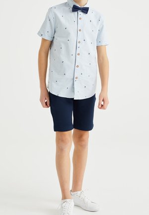 Chłopiec ubrany w jasnoniebieską koszulę z krótkim rękawem z małymi ciemnymi wzorami, granatową muchę, granatowe szorty i białe trampki, stojący.