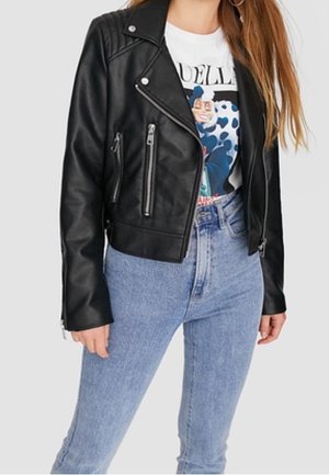 Woman wearing black leather zip-up jacket, white graphic t-shirt with cartoon design, and high-waisted light blue jeans against gray background.