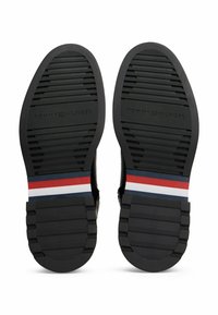 Ein Paar schwarze Schuhsohlen mit strukturierten Gripps, die rote, weiße und blaue Streifen nahe der Ferse und die Prägung "Tommy Hilfiger" aufweisen.