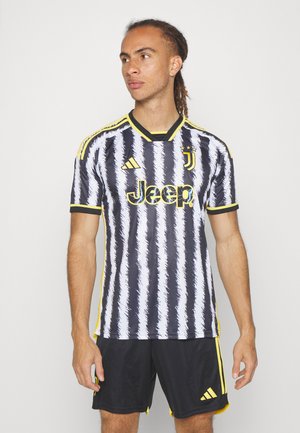 Maglia della Juventus in strisce verticali bianche e nere con dettagli gialli, caratterizzata da un collo a V e un logo, indossata con pantaloni corti neri.