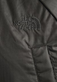 Mörkgrå ripstoptyg med svart broderad "THE NORTH FACE"-logotyp och en sydd rektangulär patchdetalj.