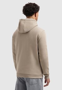Persoon met krullend haar gezien van achteren, gekleed in een beige hoodie en lichtblauwe spijkerbroek tegen een effen achtergrond.