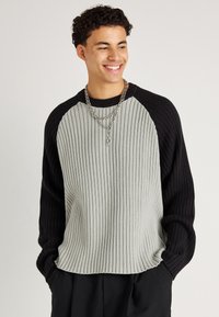 Grau und schwarz gestreifter Strickpullover mit Rundhalsausschnitt, der kontrastierende Raglanärmel und einen lockeren Schnitt aufweist. Silverner geschichteter Kettenanhänger.